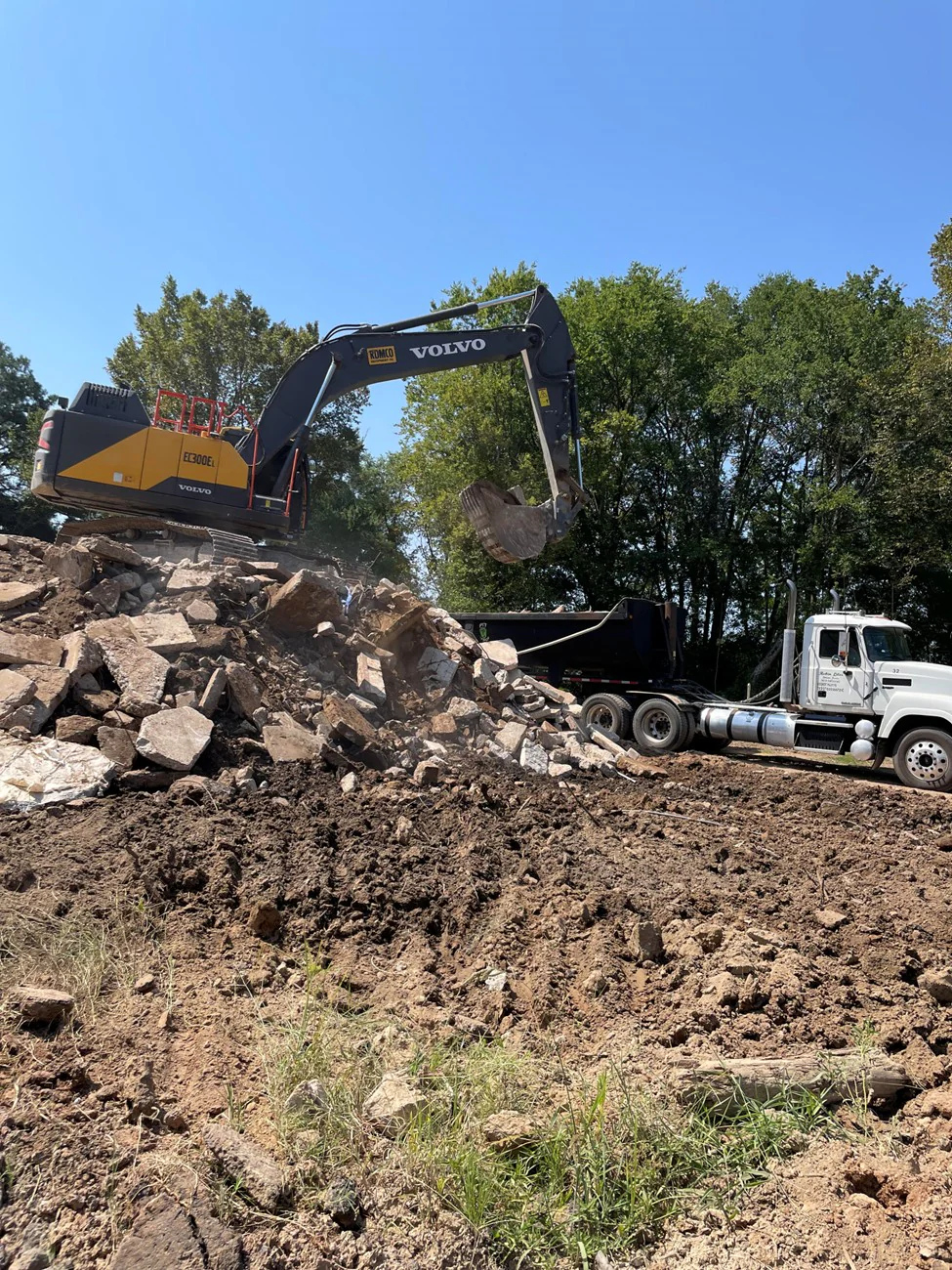 Excavator breaking down concrete beside a haul truck.